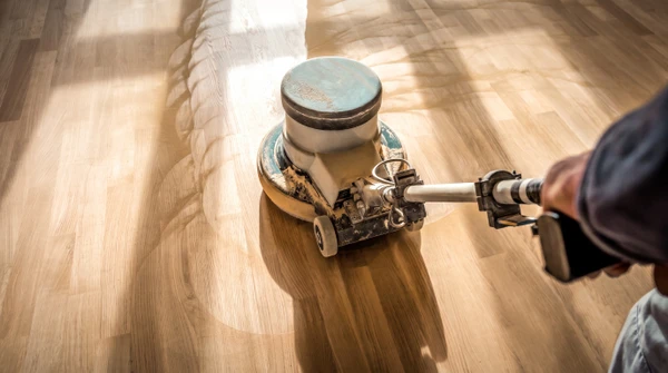 Gros plan d'une ponceuse professionnelle équipée d'un système d'extraction de poussière (tube flexible et collecteur) en action sur une surface de parquet massif. Perspective dynamique montrant le plateau de ponçage en contact avec les lames de bois, la poussière capturée par le système d'aspiration, et le détail du grain du parquet soulevé par l'abrasif. Mains gantées du professionnel guidant l'équipement. Éclairage naturel révélant la finesse de la poussière filtrée et l'équipement de pointe. Pas de logo ni texte visible.